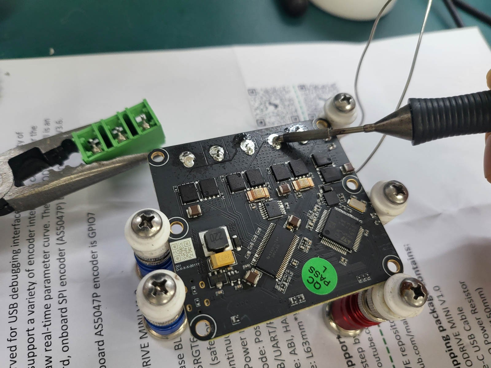 Back of the XDrive MINI driver board held in a magnetic PCB holder. A soldering iron heats the terminal block's solder joints while long-nose pliers hold the removed terminal block.