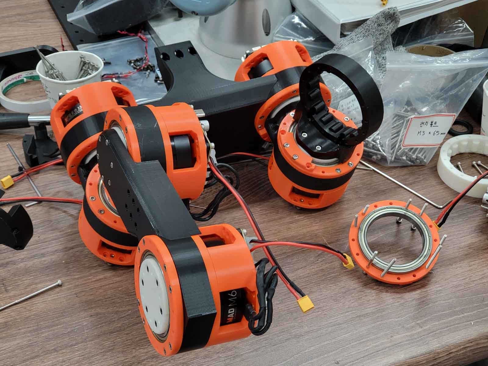 A leg being assembled on a workbench: several orange and black actuators joined by black leg structure parts, with bolts and an actuator ring nearby.
